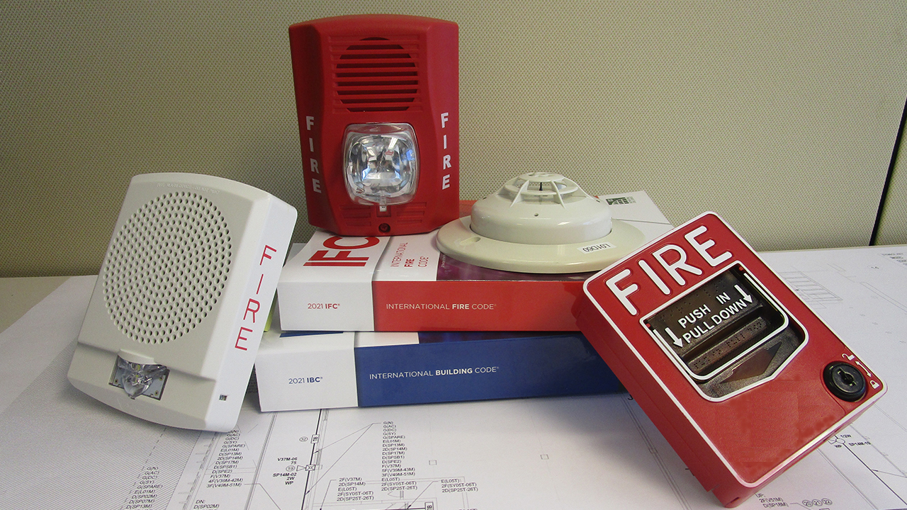 Fire alarms with speakers and strobes, a pull station, and code books stacked on a set of blue print. and code books